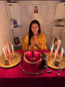 Melissa de vestido amarelo rezando de olhos fechados diante de uma mesa vermelha com velas, pétalas e a imagem de Santo Antônio
