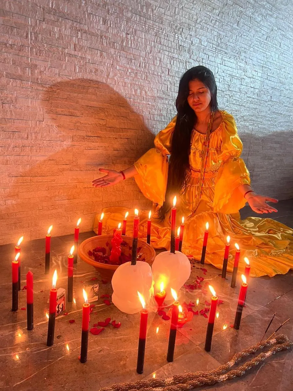 Melissa de vestido amarelo, de olhos fechados e braços abertos, atrás de um círculo de velas vermelhas e pretas